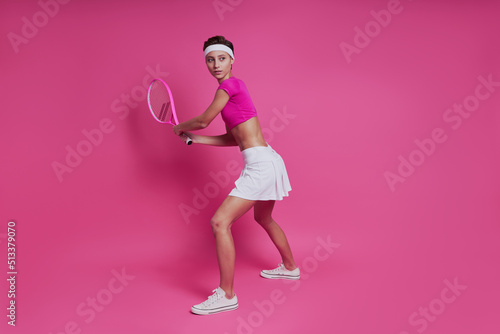 Attractive young woman holding tennis racket while standing against pink background © gstockstudio