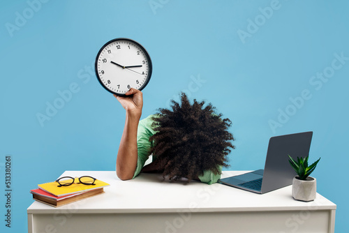 Tired black male student lying at desk with pc laptop and books, holding clock and putting head on hand, blue background