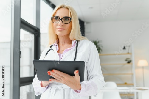 Portrait of beautiful mature woman doctor holding digital tablet. Confident female doctor using digital tablet with colleague talking in background at hospital.