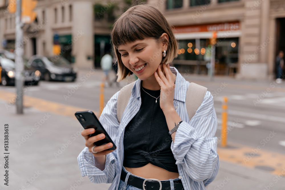 Adorable smiling straight short hair smiling while holding smartphone in the city wearing blue shirt and black top. Outside phot of caucasian woman wallking in city in warm day with backpack Adorable smiling straight short hair smiling while holding smartphone in the city wearing blue shirt and black top. Outside phot of caucasian woman wallking in city in warm day with backpack
