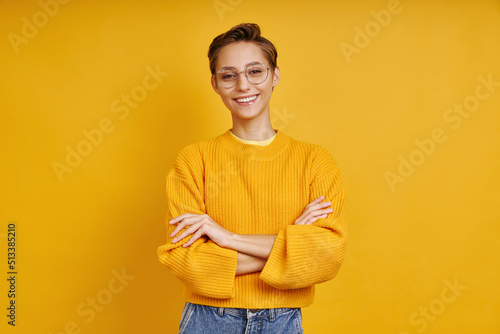 Beautiful young woman keeping arms crossed while standing against yellow background