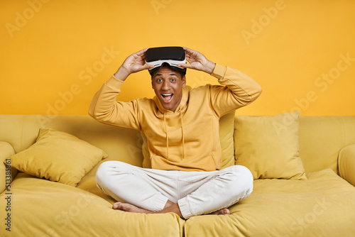 Happy young man wearing virtual reality glasses while relaxing on the couch with yellow background © gstockstudio
