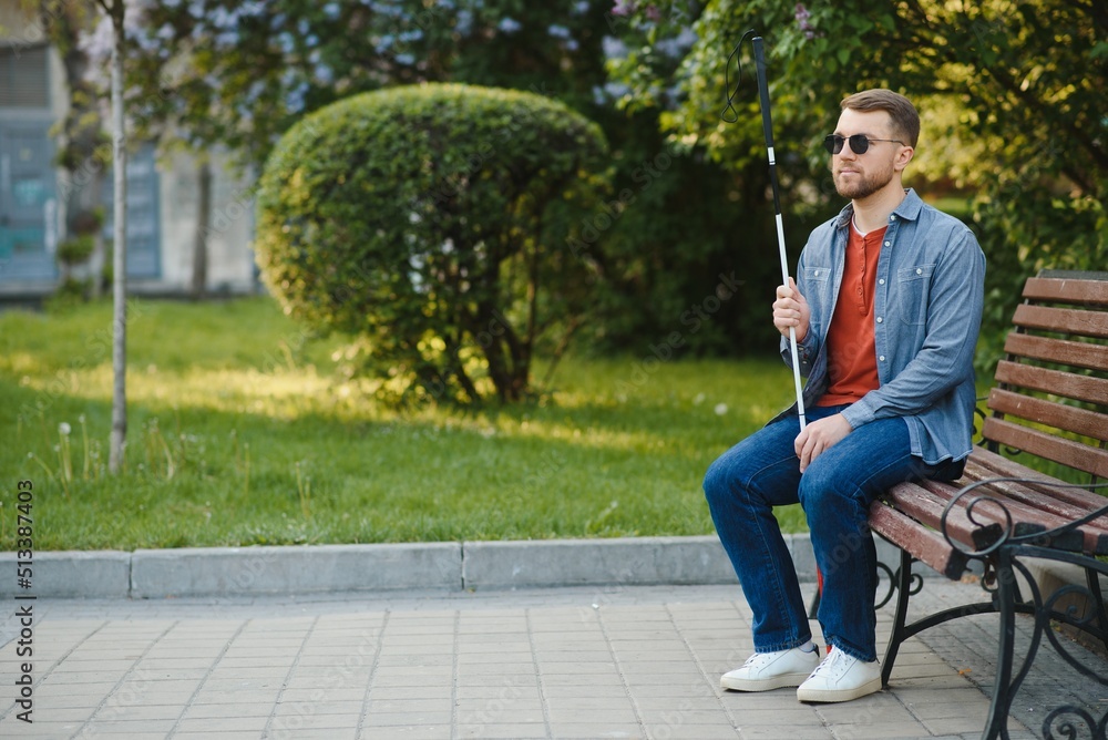 Obraz premium Visually impaired man with walking stick, sitting on bench in city park. Copy space