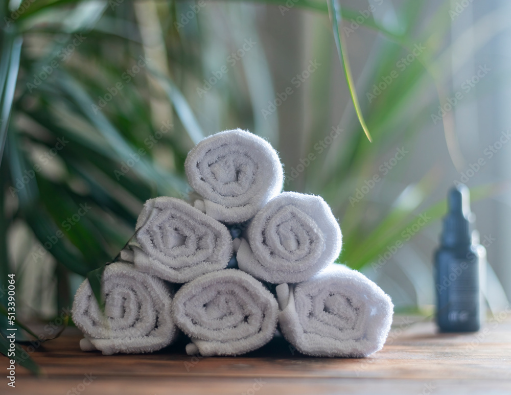 Fototapeta premium white rolled cotton towels stacked in a pyramid on a wooden table against a background of green plants and an oil bottle.