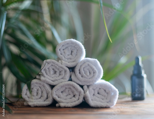white rolled cotton towels stacked in a pyramid on a wooden table against a background of green plants and an oil bottle.