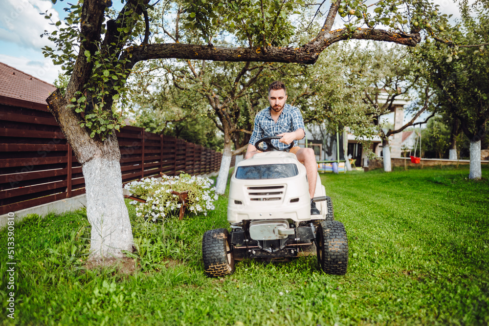 Fototapeta premium Gardener using lawn tractor for mowing grass in garden. Landscaping works with professional tools
