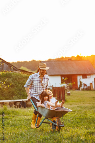 Happy little children having fun in a wheelbarrow pushing by dad in domestic garden on warm sunny day. Active outdoors games for family with kids. Spending time together on weekend at the countryside.