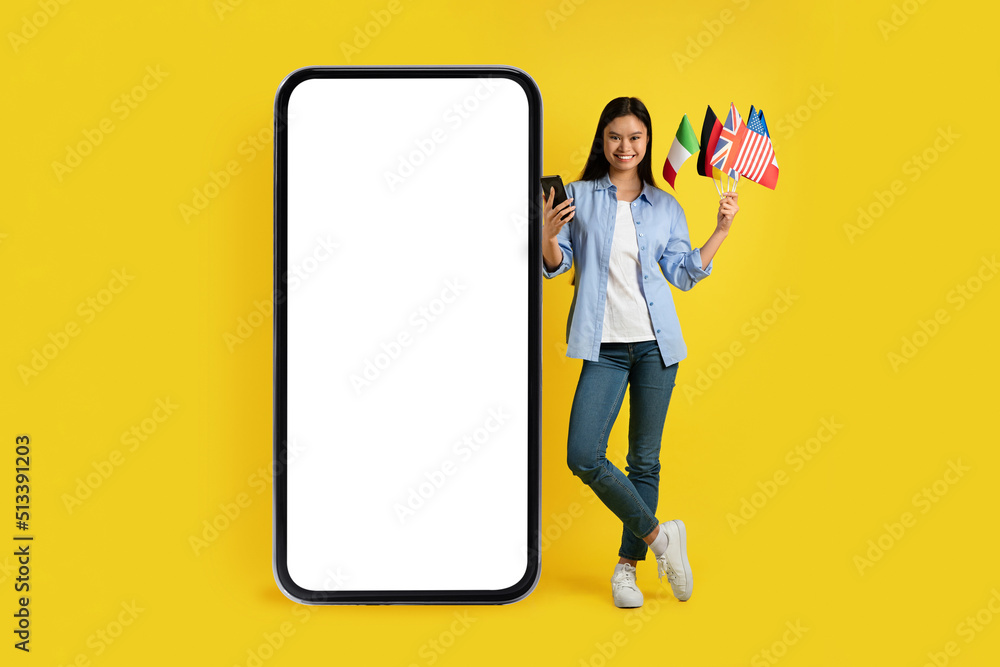Smiling young asian lady with smartphone and many international flags standing near huge phone with blank screen Smiling young asian lady with smartphone and many international flags standing near huge phone with blank screen