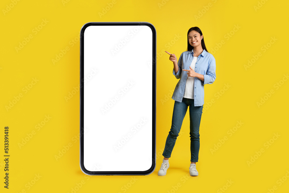Social networks and chat. Cheerful happy young asian lady pointing finger at huge phone with blank screen Social networks and chat. Cheerful happy young asian lady pointing finger at huge phone with blank screen