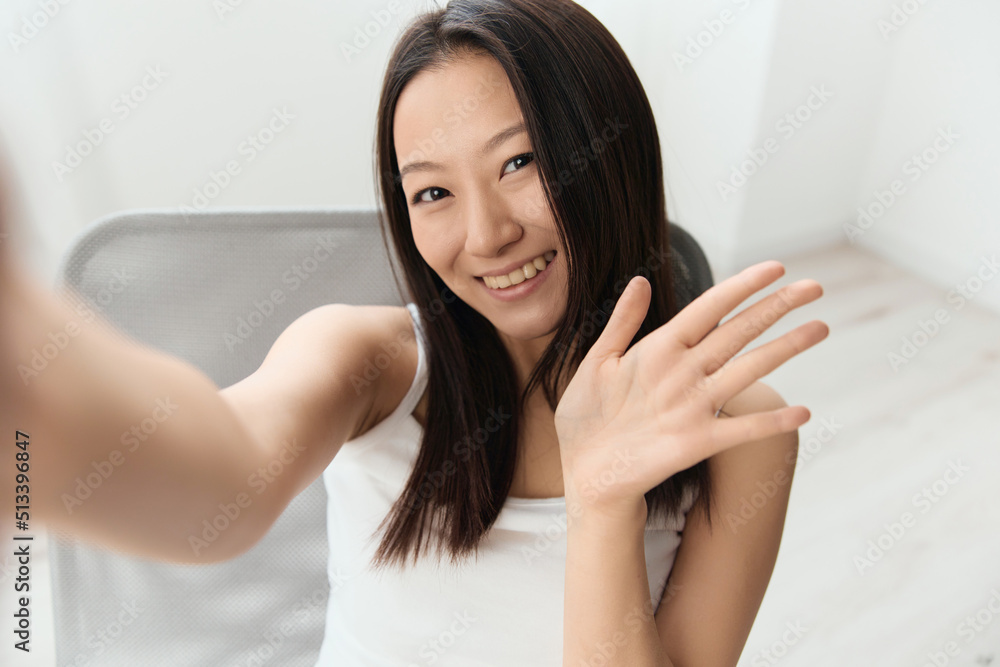 Friendly happy tanned lovely young Asian woman waving hand greeting at camera doing selfie at home interior living room. Distance communication Social media concept. Cool offer Wide angle Selfie view Friendly happy tanned lovely young Asian woman waving hand greeting at camera doing selfie at home interior living room. Distance communication Social media concept. Cool offer Wide angle Selfie view