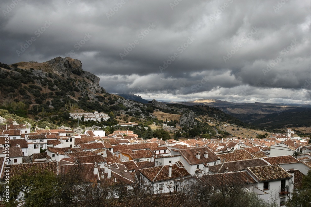 Obraz premium Red terracotta roofs of the white village of Grazalema, Spain, against a backdrop of spectacular dark storm clouds and the dry autumn slopes of yellow brown grass of the Sierra del Pinar mountains