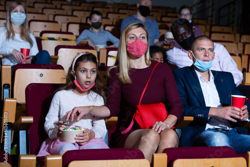 Interested parents with tween daughter wearing protective masks watching movie in cinema. Concept of family leisure in coronavirus pandemic © JackF