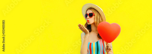 Portrait of beautiful young woman with big red heart shaped balloon blowing her lips wearing summer straw hat, sunglasses on yellow background, blank copy space for advertising text