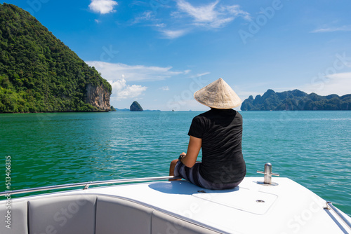 young man in a Vietnamese hat sits on the prow of a boat in a lotus position against the backdrop of limestone cliffs in the Andaman Sea, Thailand