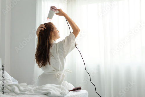 Foto Young Asian woman in bathrobe uses hair dryer on bed.