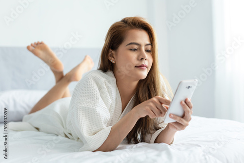 Smiling Asian woman lying down the bed while using smartphone with her legs raised slightly at home bedroom.