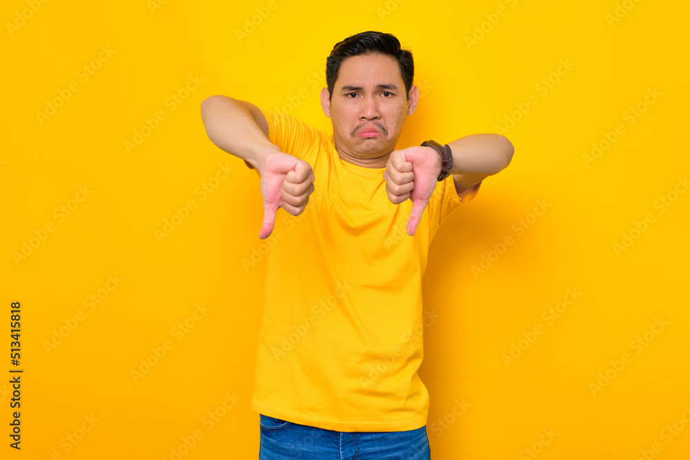 Upset young Asian man in casual t-shirt showing thumb down with expressing disapproval isolated on yellow background. People lifestyle concept Upset young Asian man in casual t-shirt showing thumb down with expressing disapproval isolated on yellow background. People lifestyle concept