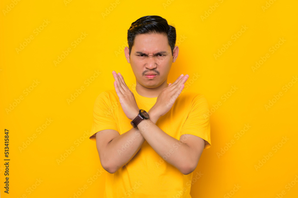 Annoyed young Asian man in casual t-shirt crossing hands making stop gesture and demonstrates rejection isolated on yellow background. People lifestyle concept Annoyed young Asian man in casual t-shirt crossing hands making stop gesture and demonstrates rejection isolated on yellow background. People lifestyle concept