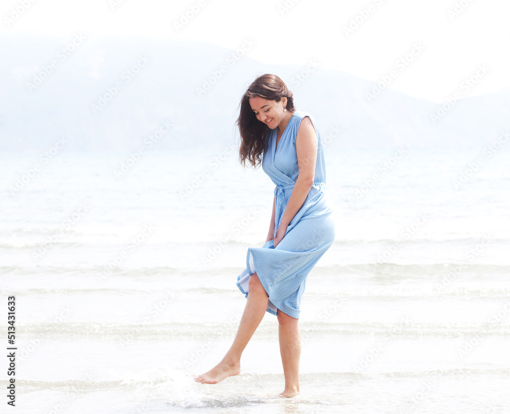Young brunette woman in a blue dress walking barefoot on a beach and dangles his feet in the water. Young brunette woman in a blue dress walking barefoot on a beach and dangles his feet in the water.