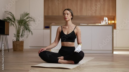 Young disabled woman is meditating and sitting on floor in apartment interior spbas. 4k Beautiful female with bionic prosthesis closes eyes and meditates, sits in lotus position on mat in bright room photo