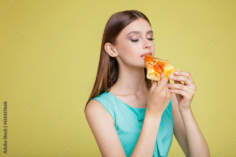 Beautiful young happy woman in a pretty aqua blue dress with  posing on yellow  background. Slim figure, studio shot. Delicious slice of pizza in hands. Model eats pizza