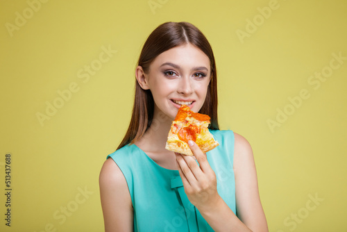 Beautiful young happy woman in a pretty aqua blue dress with posing on yellow background. Slim figure, studio shot. Delicious slice of pizza in hands. Model eats pizza