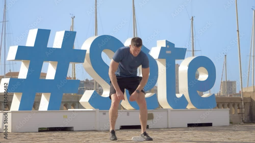 a young athletic Caucasian man turning on his music speaker to do squats in front of the hastag of the city of Sète (south of France)