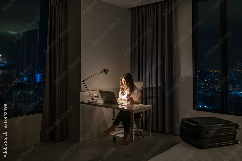 Work and travel. Young Asian traveler using laptop on table at the lamp with making video call meeting to team online in hotel room at night during a summer vacation holiday travel and suitcase on bed
