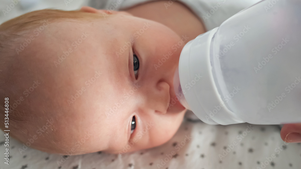 Newborn baby girl with half closed eyes drinks water from plastic bottle lying on bed. Mother feeds cute little kid at home closeup, side view Newborn baby girl with half closed eyes drinks water from plastic bottle lying on bed. Mother feeds cute little kid at home closeup, side view