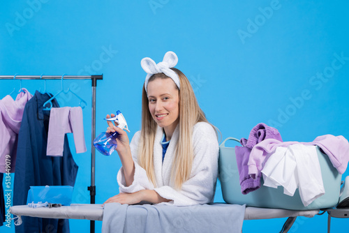 A smiling blonde in a bathrobe is cleaning clothes in the morning  ironing  holding a spray bottle of water. Portrait of housewife on blue background in studio.
