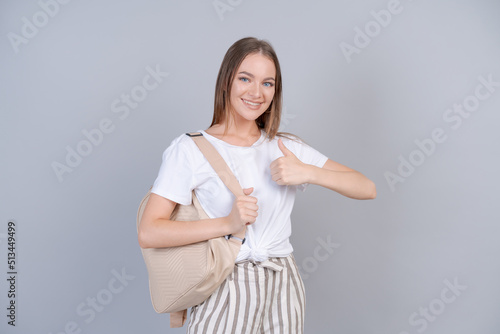 A beautiful girl in white t-shirt with backpack on her back, student smiles cutely on a gray background in the studio, a happy young woman rejoices at the day off. Advertising and mockup