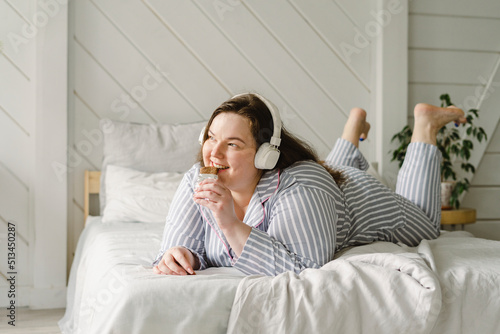 Happy woman eating chocolate and listening music through wireless headphones lying on bed at home photo