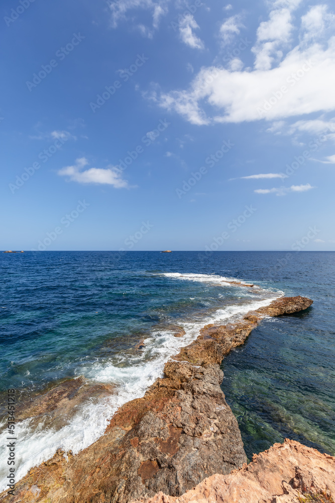 Vertical shot: narrow strip of rock gently descends into sea waters and is lost somewhere in distance, leaving foam from sea waves, Ibiza, Balearic Islands, Spain