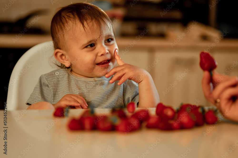 a happy baby looks happily at a strawberry in his dad's hand, the child is at home in the kitchen eating a berry a happy baby looks happily at a strawberry in his dad's hand, the child is at home in the kitchen eating a berry
