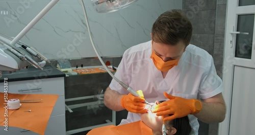doctor dentist man in mask and gloves with professional equipment cleans teeth patient from plaque, client with mouth open with dilator lies in chair under bright lighting