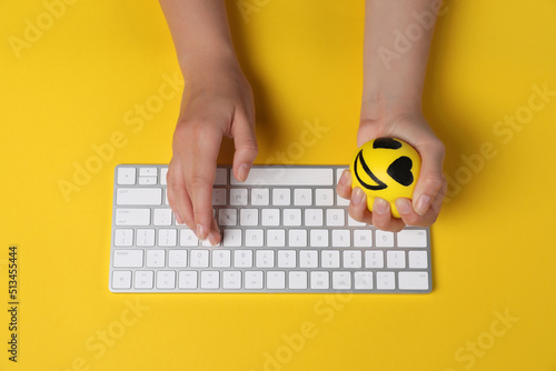 Woman squeezing antistress ball while typing on keyboard against yellow background, top view photo