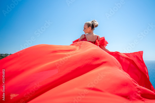 Blonde with long hair on a sunny seashore in a red flowing dress  back view  silk fabric waving in the wind. Against the backdrop of the blue sky and mountains on the seashore.