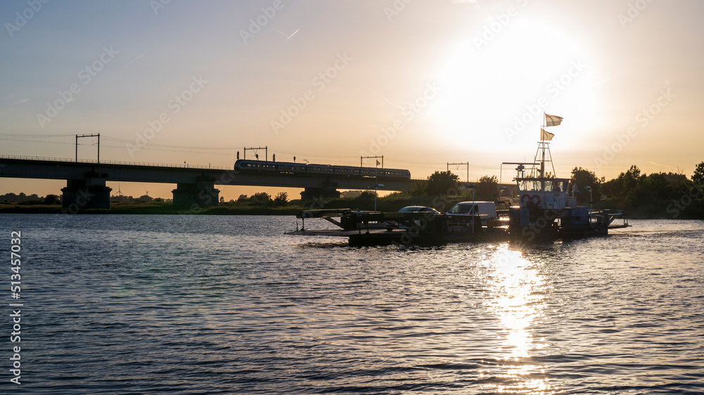 Silhouette of ferry boat, train and bridge early evening. Sunshine reflecting in the river. It is the ferry over the river Lek, at Culemborg, the Netherlands.