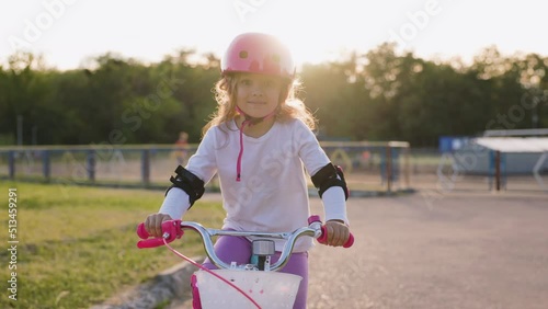 Cute light hair little girl in pink helmet in elbow and knee pads rides a bicycle at the street. Sunset background. Sport activity for children. Medium shot photo