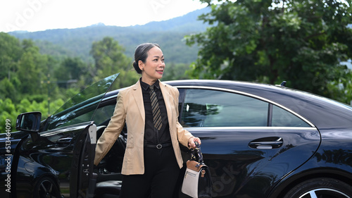 Elegant mature businesswoman standing by open door of black car and looking alway