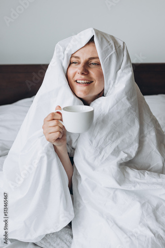 Young happy redhead woman with cup of coffee sitting in bed under blanket at home bedroom. comfort or home weeekend concept, mockup of mug photo