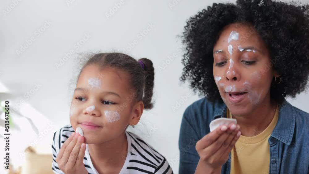 Close view of mother, daughter are applying cream on face and standing in home room spbi. 4k American african woman, girl spread lotion on skin and look with smiles, stand in light interior. Modern Close view of mother, daughter are applying cream on face and standing in home room spbi. 4k American african woman, girl spread lotion on skin and look with smiles, stand in light interior. Modern