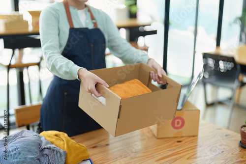 Portrait of a small startup Asian female entrepreneur SME owner picking up a yellow shirt before packing it in an inner box with a customer. Online Business Ideas and Freelance