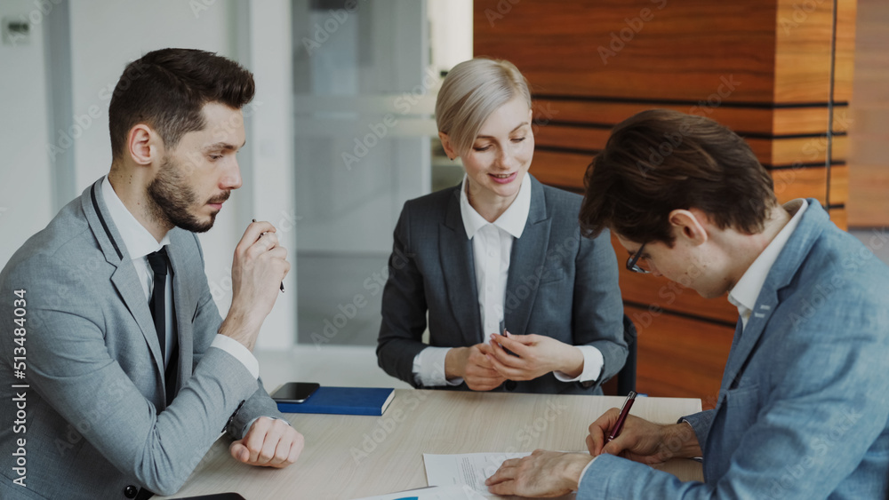 Fototapeta premium Young businessman in suit signing contract with his colleague while corporate lawyer explaining details of partnership sitting in modern office indoors