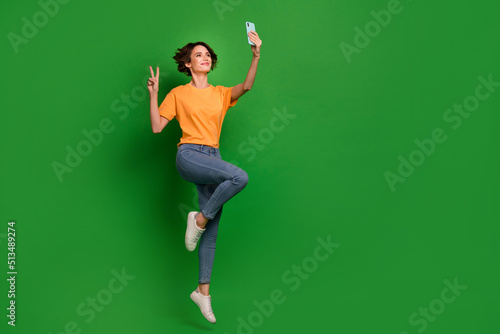 Full size photo of excited sporty girl jump hold telephone take selfie show v-sign isolated on green color background