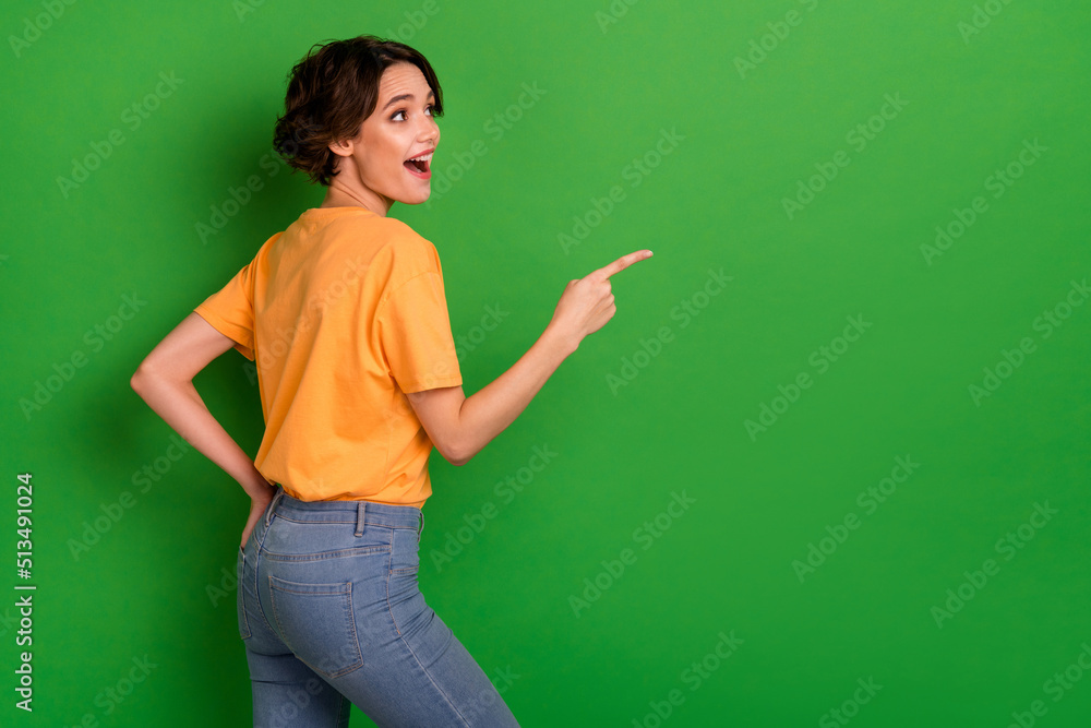 Photo of cute excited young woman dressed orange t-shirt looking pointing empty space isolated green color background