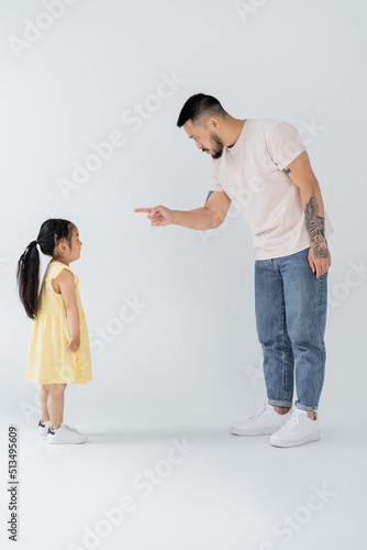 full length of asian father pointing at preschooler daughter while punishing her on grey.