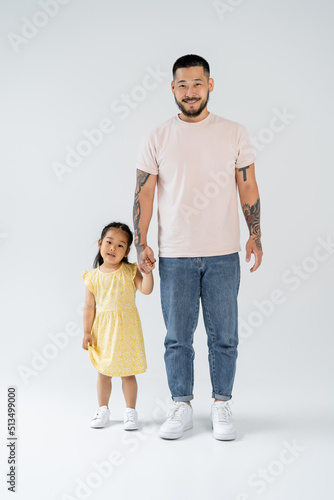 full length of happy and tattooed asian father holding hands with preschooler daughter on grey.