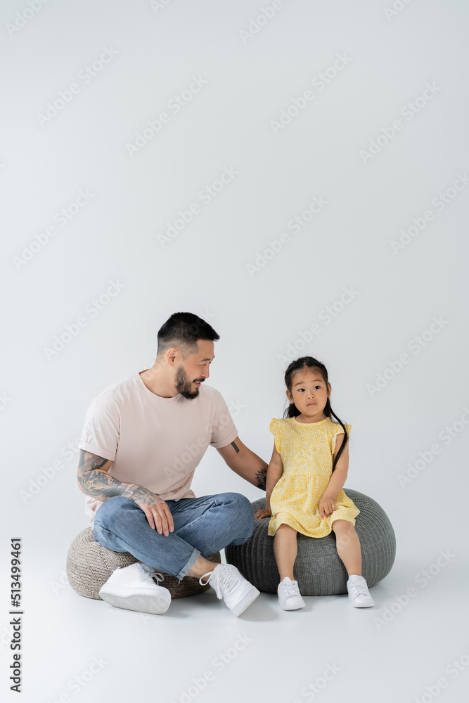 full length of happy tattooed father and asian daughter in yellow dress sitting on puff chairs on grey. full length of happy tattooed father and asian daughter in yellow dress sitting on puff chairs on grey.