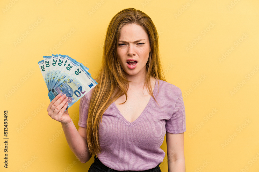 Young caucasian woman holding banknotes isolated on yellow background screaming very angry and aggressive. Young caucasian woman holding banknotes isolated on yellow background screaming very angry and aggressive.
